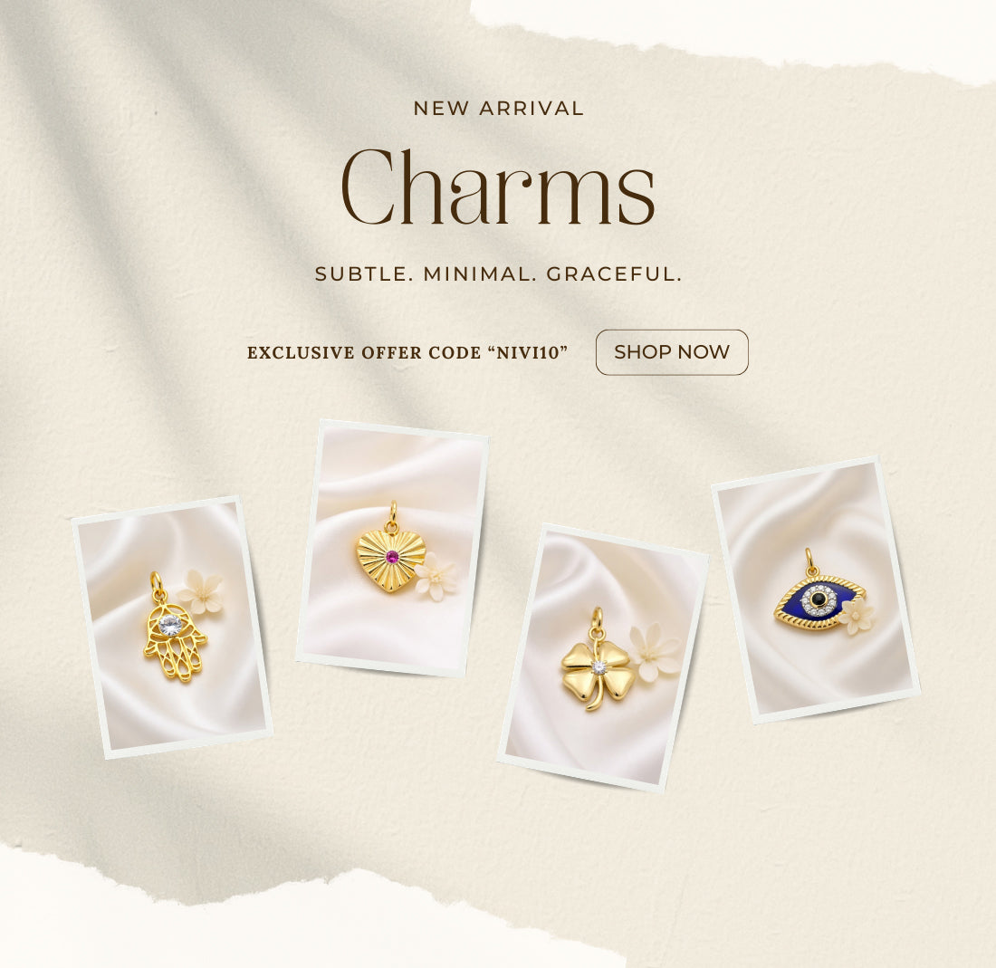 Homepage banner showcasing NIVI's Charms Collection featuring four handcrafted charm pendants displayed on elegant satin-inspired cards against a soft ivory background. The collection includes a Hamsa hand charm, radiant heart charm, four-leaf clover charm, and evil eye charm, crafted in 925 sterling silver with one-micron gold plating. The banner highlights the message "New Arrival – Charms" with the tagline "Subtle. Minimal. Graceful." and features an exclusive offer code "