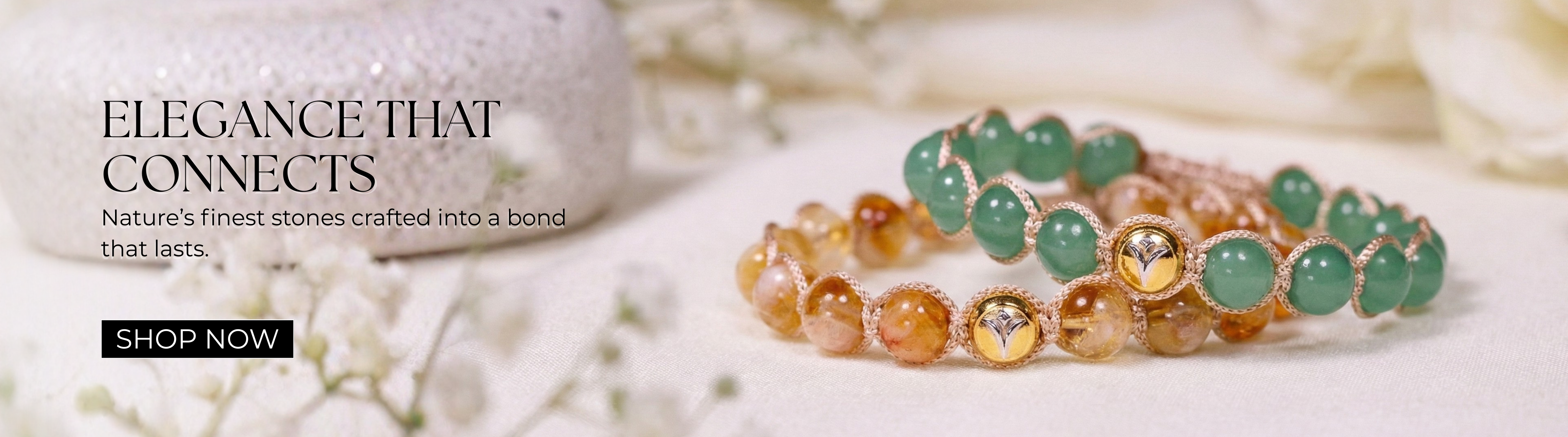 Homepage banner featuring two handcrafted natural gemstone bracelets displayed on a soft fabric background with delicate white flowers. One bracelet is crafted with polished green gemstones, while the other features warm golden and amber-toned natural gemstones. Both bracelets showcase NIVI's signature 925 sterling silver centerpiece with one-micron gold plating, highlighting timeless craftsmanship, meaningful design, and everyday elegance. The banner includes the message "Elegance That Connects – Nature's 